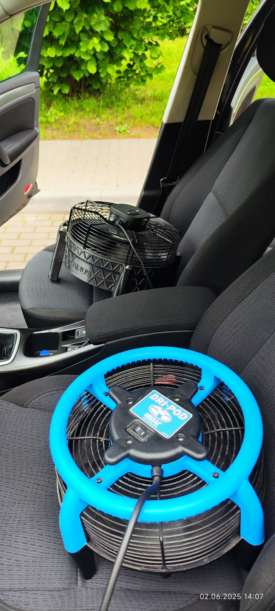 Dwa wentylatory osuszające, jeden czarny na konsoli środkowej, drugi niebieski na siedzeniu pasażera w samochodzie. Widoczne wnętrze pojazdu i fragment zieleni za oknem.