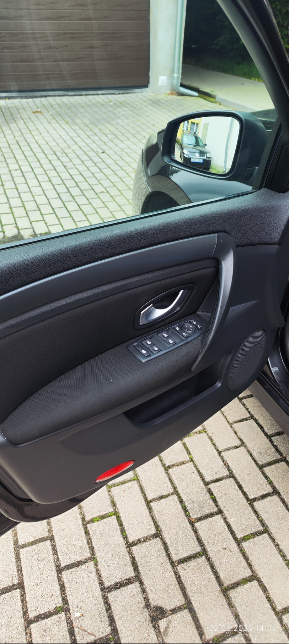 Wnętrze otwartych drzwi auta: czarna tapicerka z panelem sterowania szybami i lusterkiem wstecznym, w którym odbija się zaparkowany samochód.