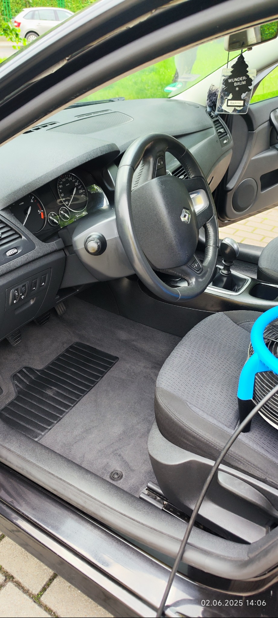 Wnętrze auta z szarą tapicerką i dywanikami, widoczny niebieski element odkurzacza. Na desce rozdzielczej zapach Wunder-Baum 'Black Ice'.