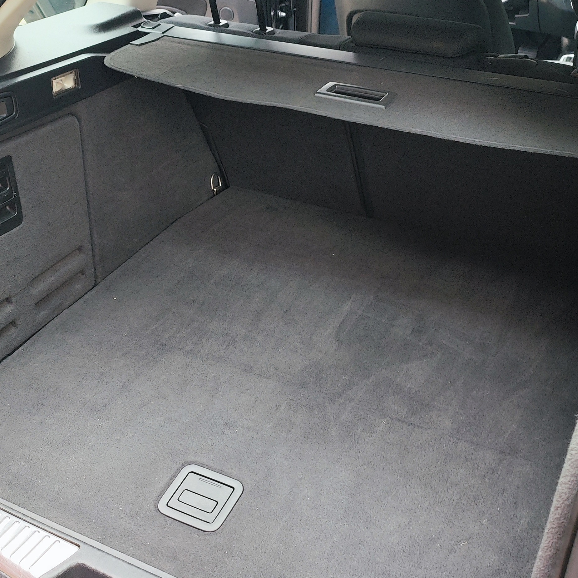 Czysty, pusty bagażnik samochodu typu kombi z szarą tapicerką i roletą. Widoczna faktura materiału i metalowe elementy mocujące.