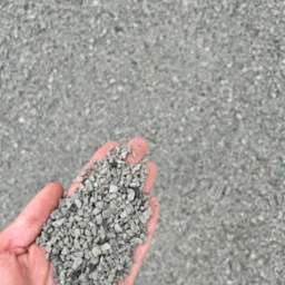 Otwarta dłoń trzymająca garść drobnego, szarego kruszywa na tle większej powierzchni tego samego materiału.