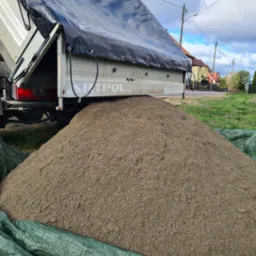 Wyładunek piasku z ciężarówki z plandeką, widoczny numer telefonu na burcie, zielona plandeka zabezpieczająca podłoże.