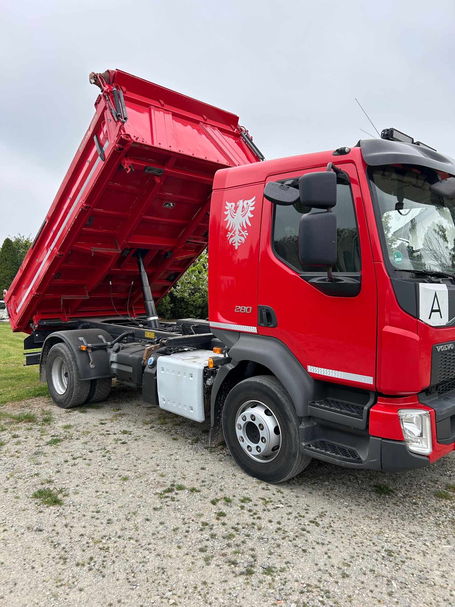 Czerwona wywrotka Volvo z podniesioną skrzynią, widoczny herb na kabinie, stojąca na żwirowym podłożu.