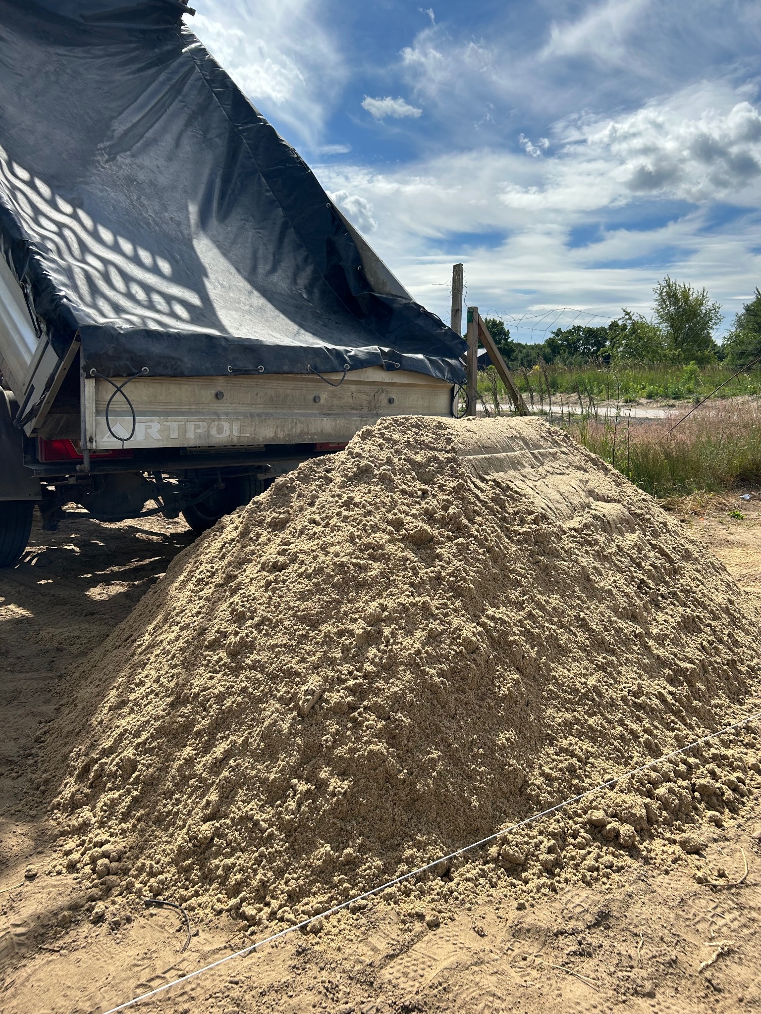 Kremowy kopiec materiału sypkiego wysypany z przyczepy ciężarowej z czarną plandeką, na tle błękitnego nieba z chmurami i zieleni za ogrodzeniem.