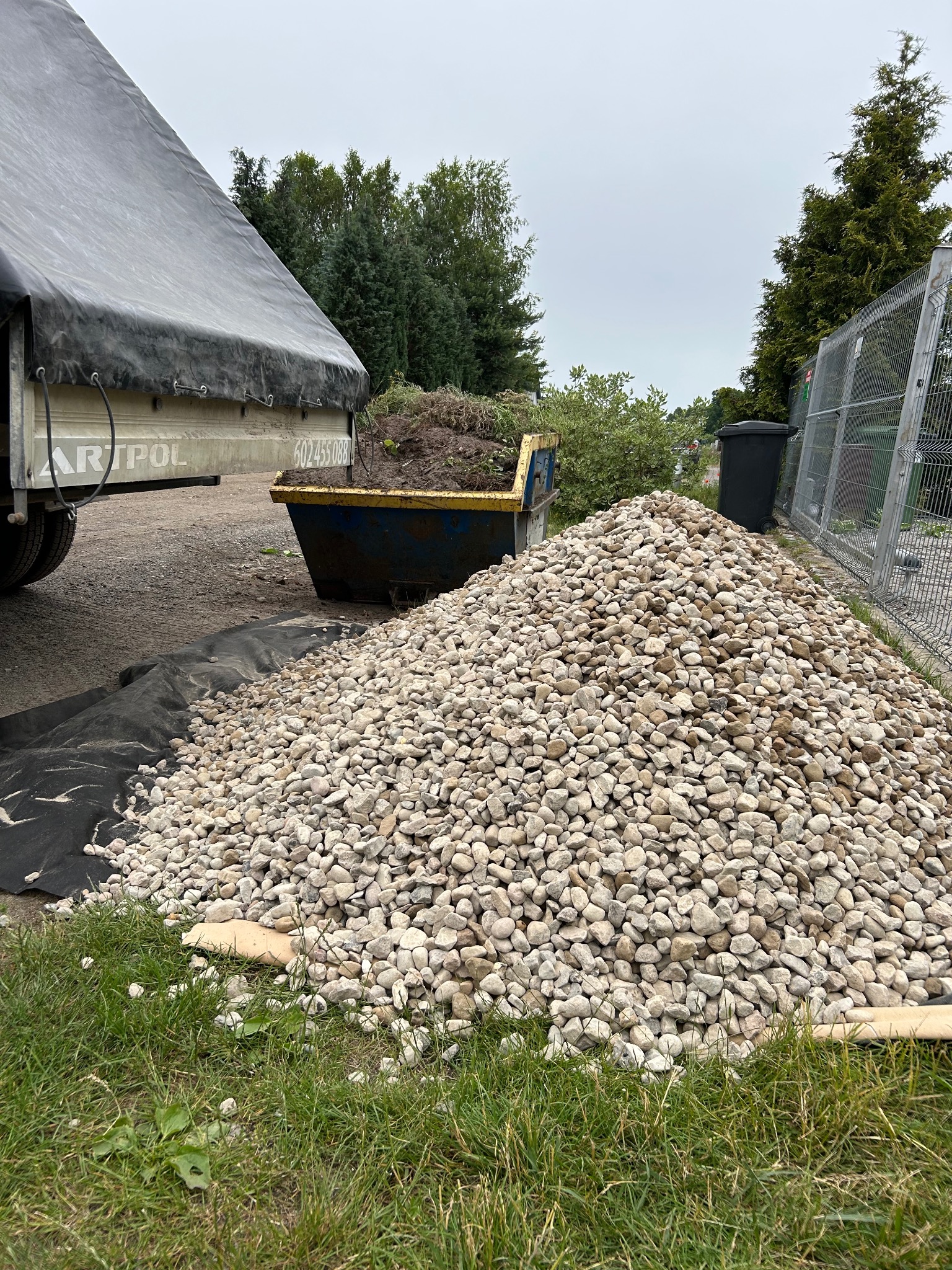 Kupa jasnych kamieni dekoracyjnych na czarnej geowłókninie, obok przyczepy z plandeką i kontenera wypełnionego ziemią, widok z poziomu gruntu.