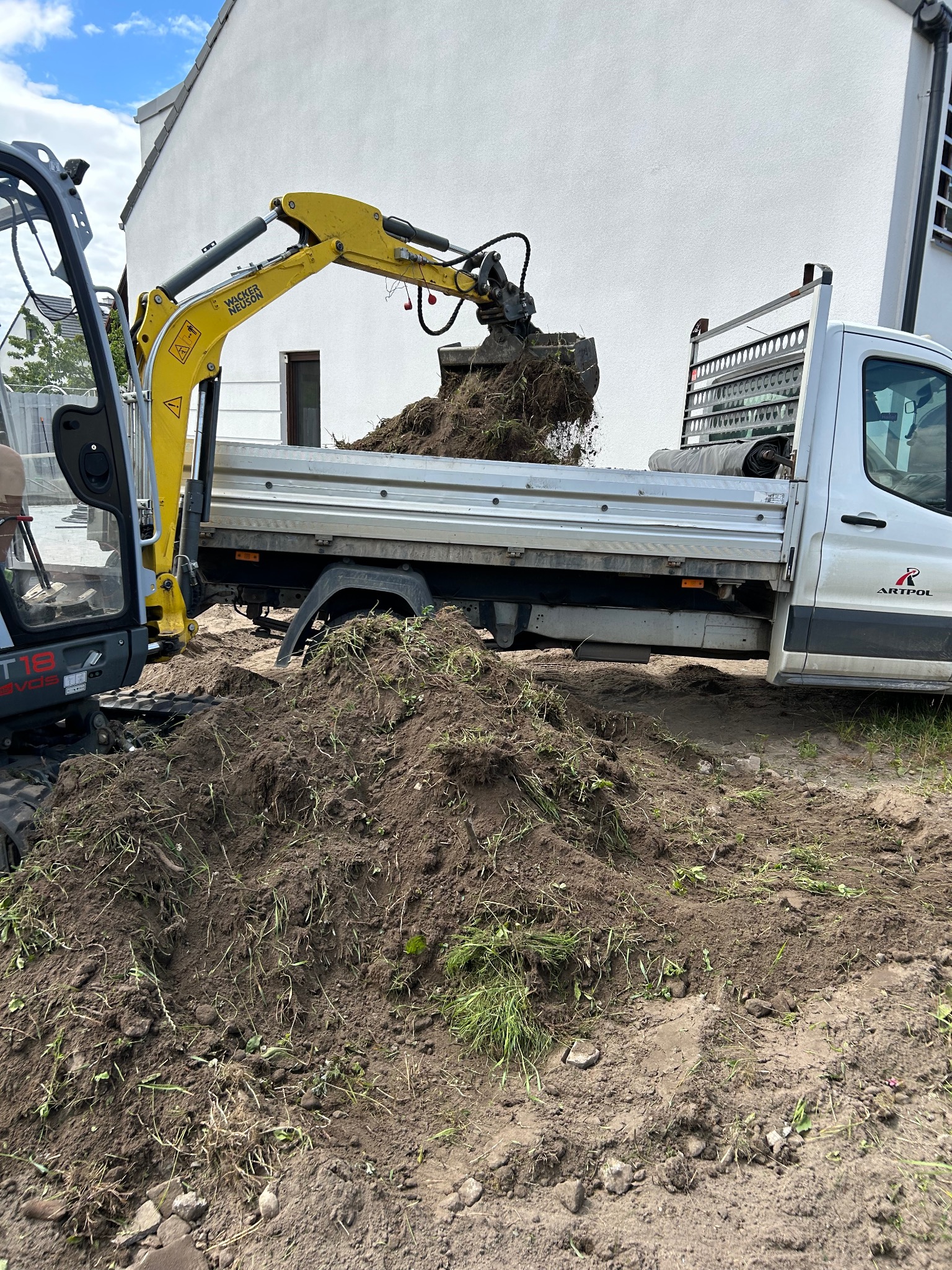 Żółta minikoparka Wacker Neuson ładuje urobek ziemny z trawą do skrzyni ładunkowej białego samochodu dostawczego Artpol na tle białego budynku.