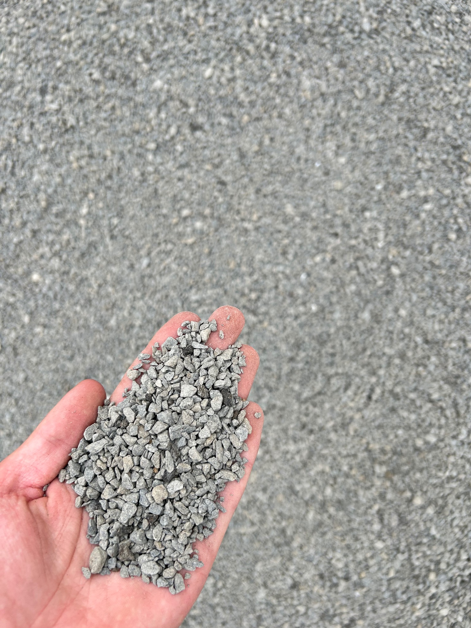 Otwarta dłoń trzymająca garść drobnego, szarego kruszywa na tle większej powierzchni tego samego materiału.