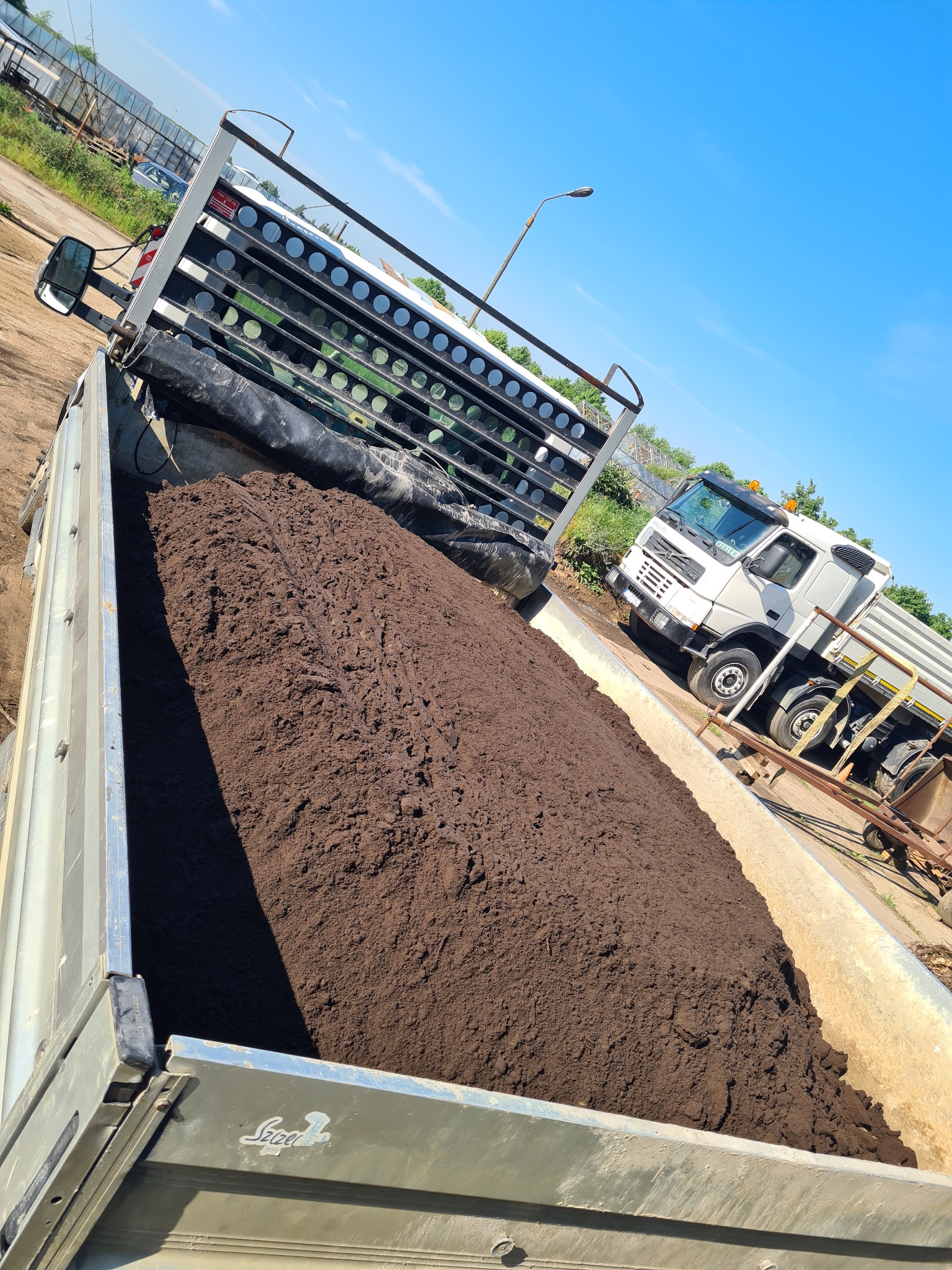 Ciężarówka z metalową skrzynią załadowana ciemną, wilgotną ziemią, widoczna z góry pod kątem, z fragmentem kabiny i otoczenia na zewnątrz.