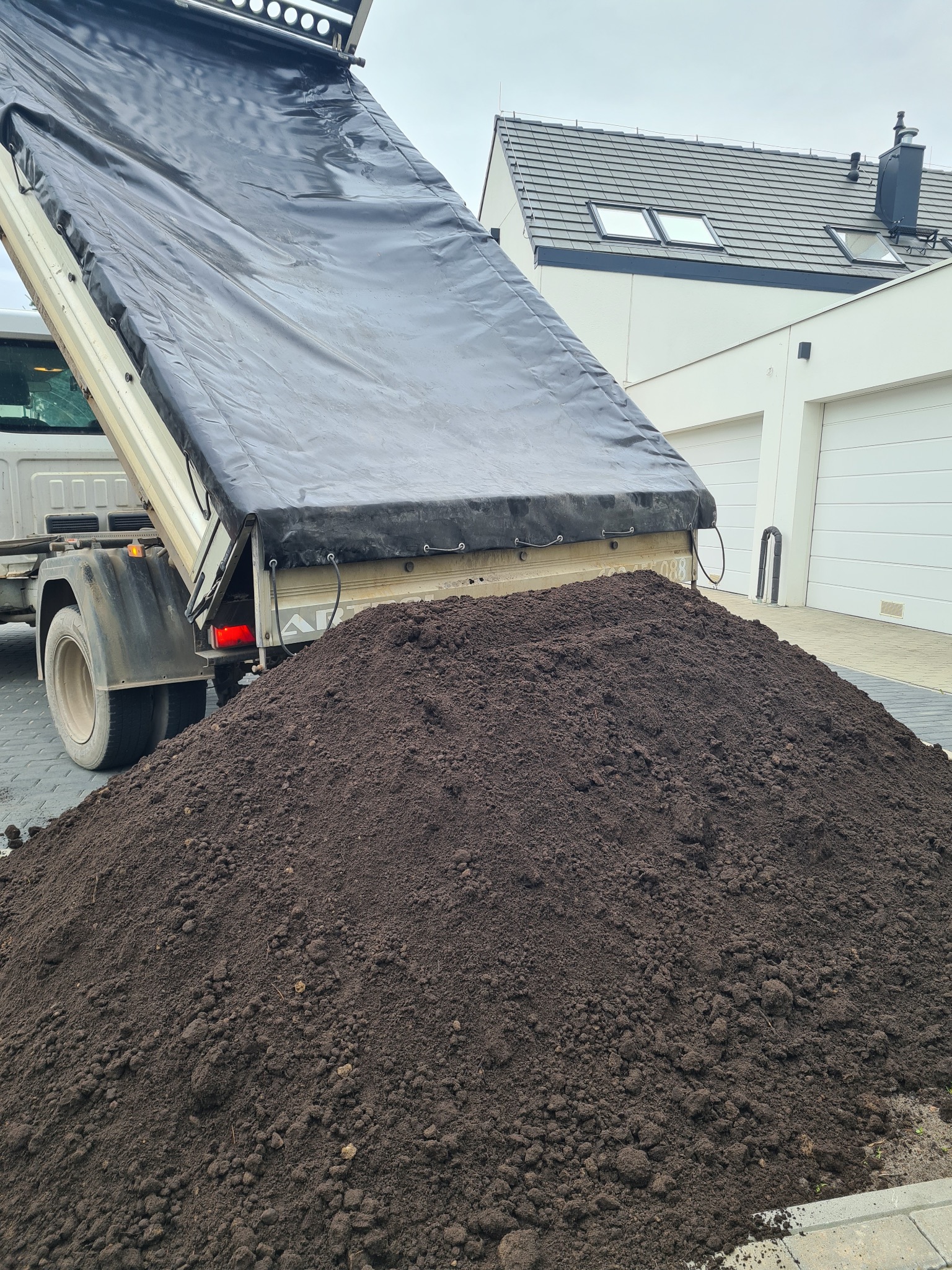 Ciężarówka z otwartą skrzynią wyładowuje dużą pryzmę ciemnej, gruzełkowatej ziemi na kostce brukowej przed nowoczesnym domem z białymi garażami.