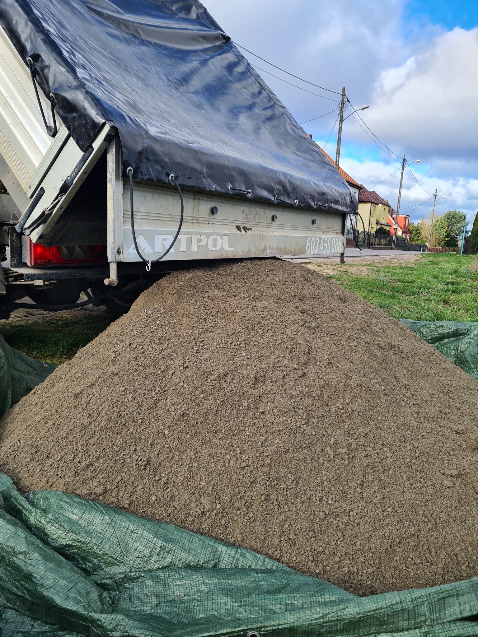 Wyładunek piasku z ciężarówki z plandeką, widoczny numer telefonu na burcie, zielona plandeka zabezpieczająca podłoże.