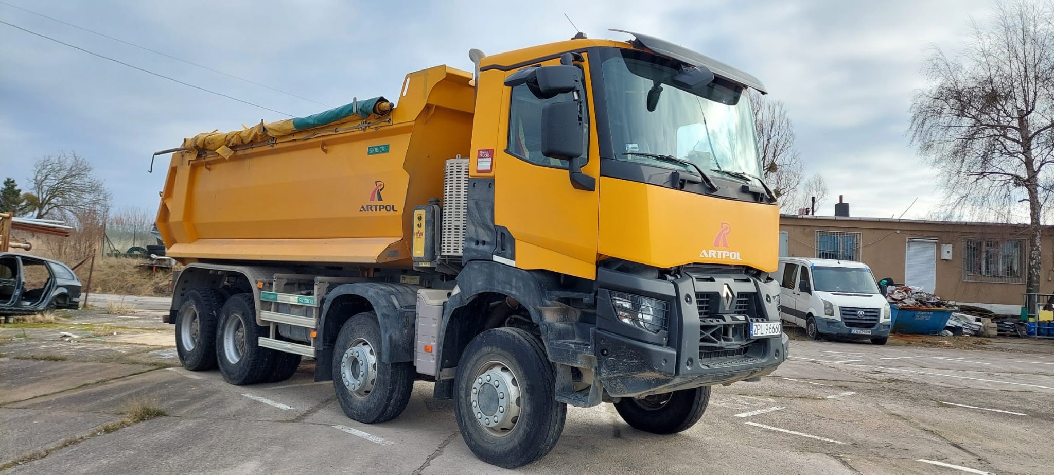 Żółta ciężarówka wywrotka marki Renault z logo firmy Artpol zaparkowana na placu, widoczna tablica rejestracyjna ZPL 96660, w tle budynek i samochód dostawczy.
