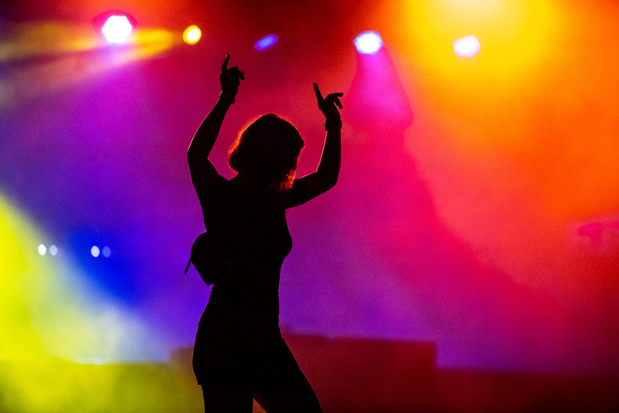Sylwetka kobiety z podniesionymi rękami na tle sceny oświetlonej kolorowymi światłami podczas koncertu. Widoczne reflektory i dynamiczne oświetlenie.