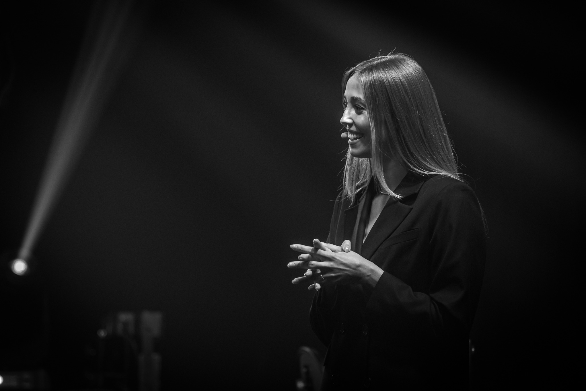 Czarno-białe zdjęcie kobiety w marynarce, z rękami złożonymi przed sobą, uśmiechającej się w świetle reflektorów na scenie podczas eventu.