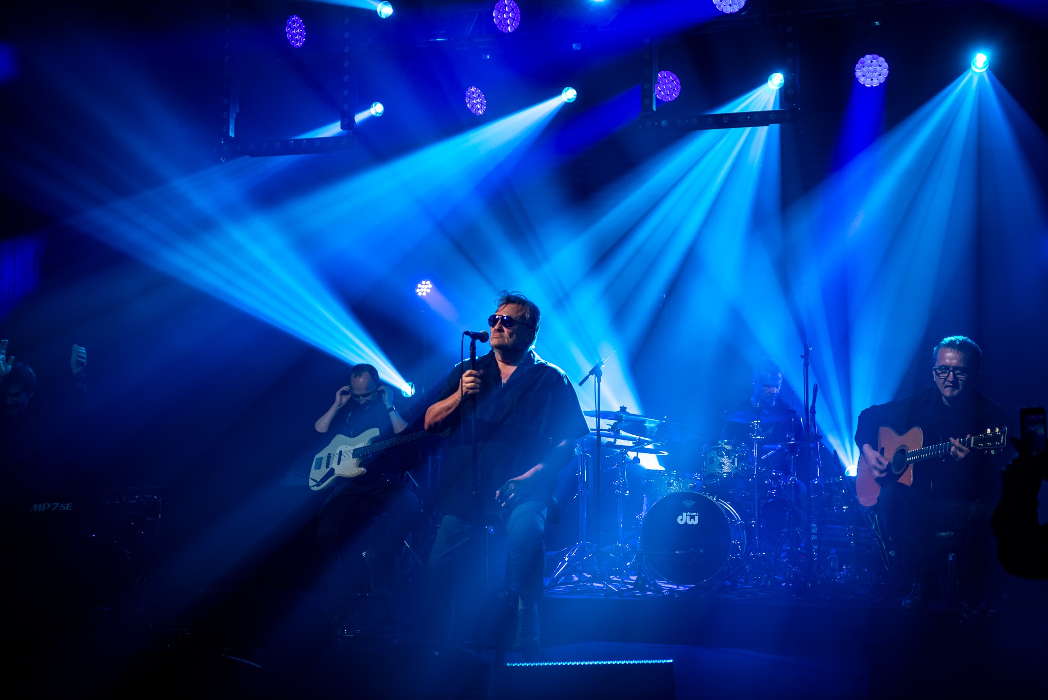 Zespół muzyczny na scenie podczas koncertu w niebieskim oświetleniu. Wokalista w okularach śpiewa do mikrofonu, a pozostali muzycy grają na instrumentach.