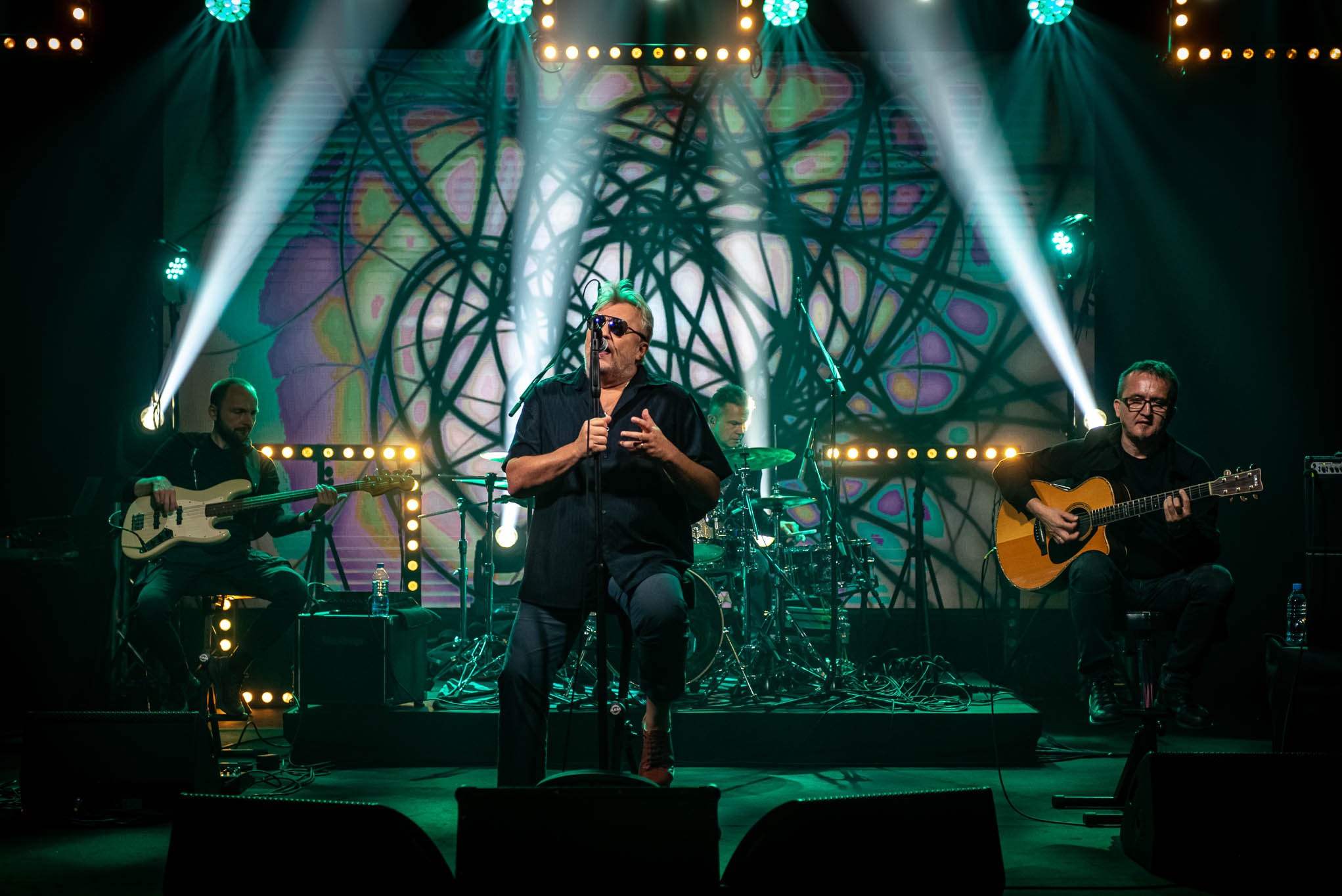 Zespół muzyczny na scenie podczas koncertu. Wokalista śpiewa do mikrofonu, gitarzysta i basista grają na instrumentach. Scena oświetlona reflektorami.