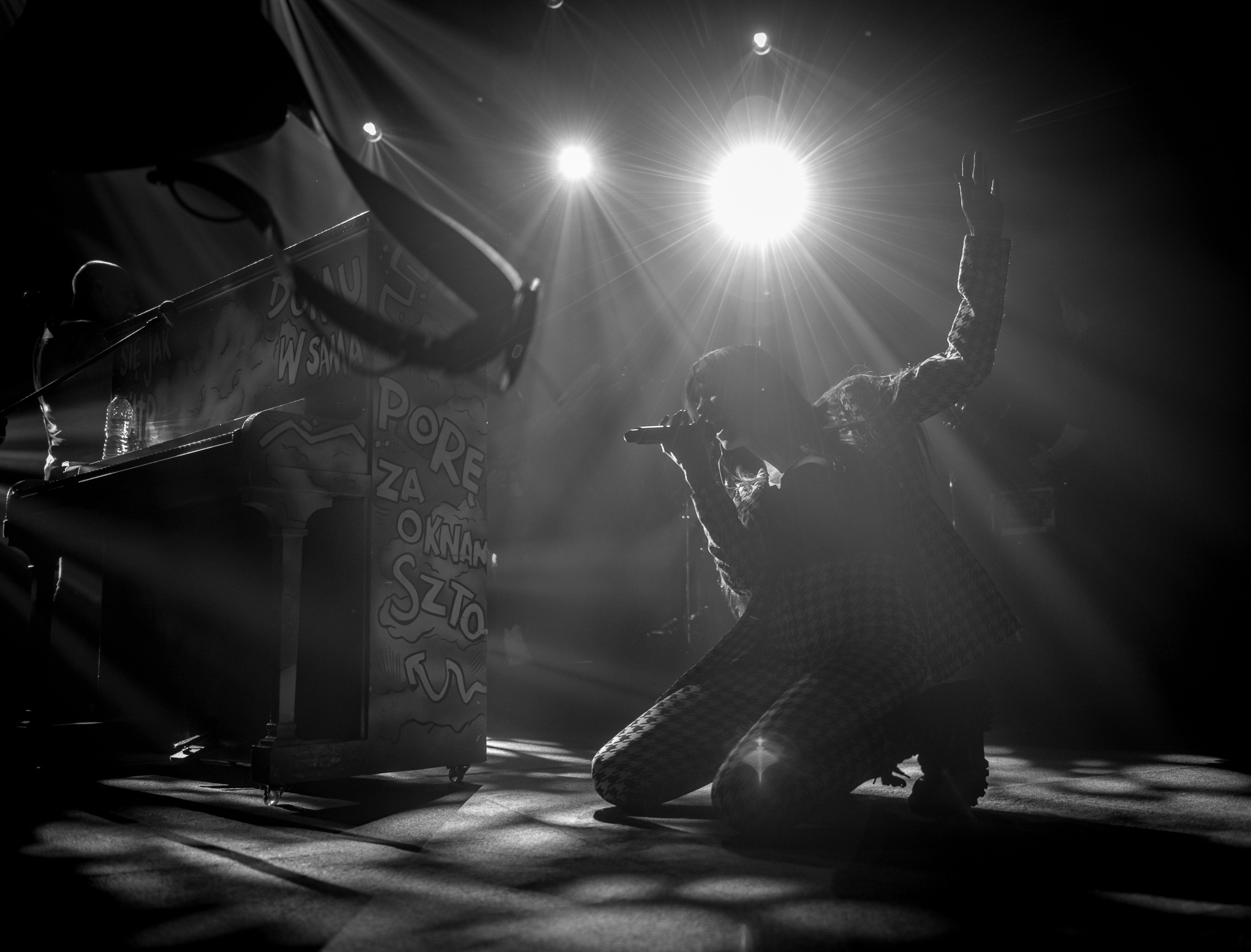 Czarno-białe ujęcie piosenkarza na kolanach, z mikrofonem, w reflektorach, obok pianino z graffiti. Dynamiczna scena koncertowa, silny kontrast, artystyczny klimat.