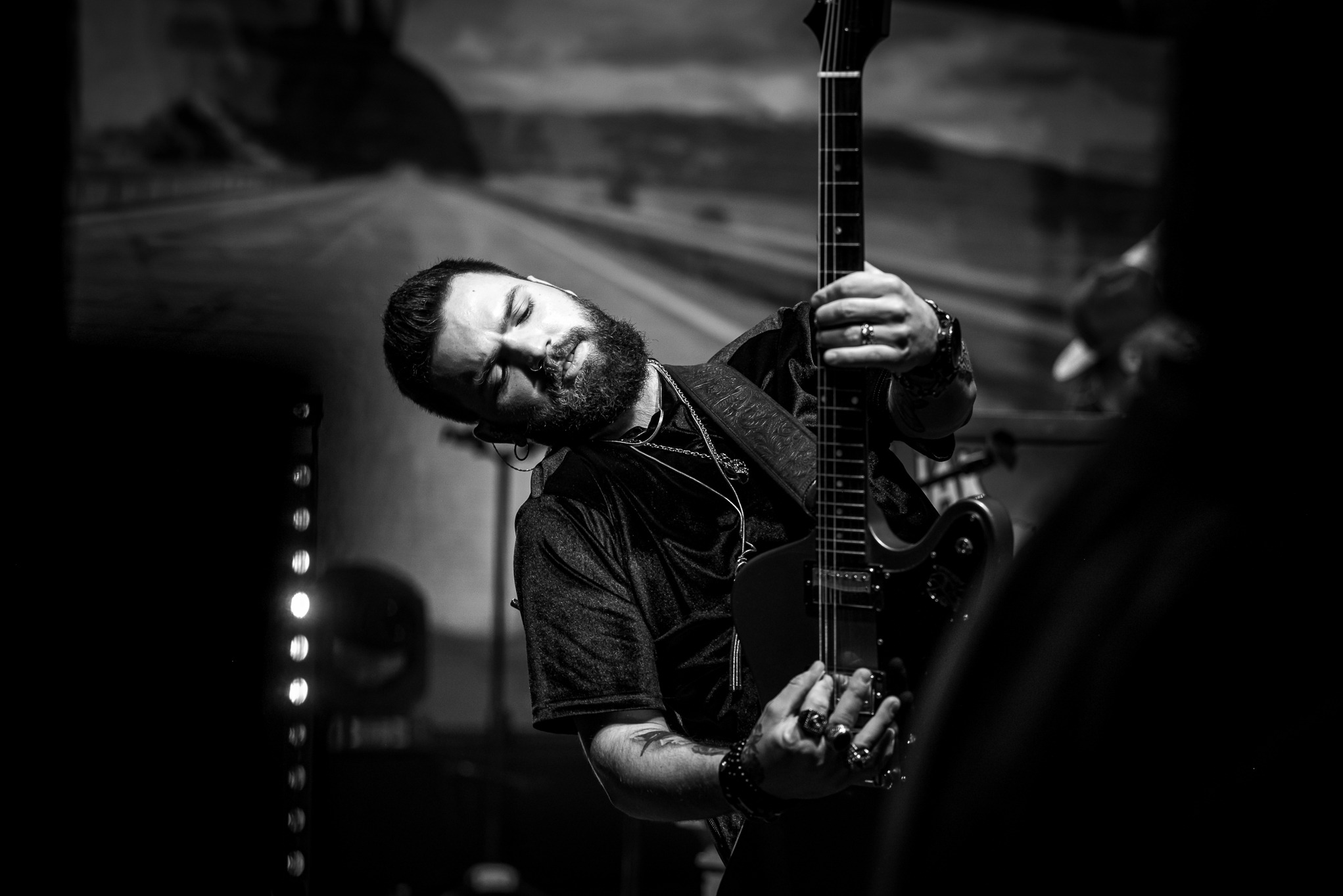 Czarno-białe zdjęcie muzyka z brodą, pochylonego z zamkniętymi oczami, trzymającego gitarę elektryczną na scenie podczas koncertu.