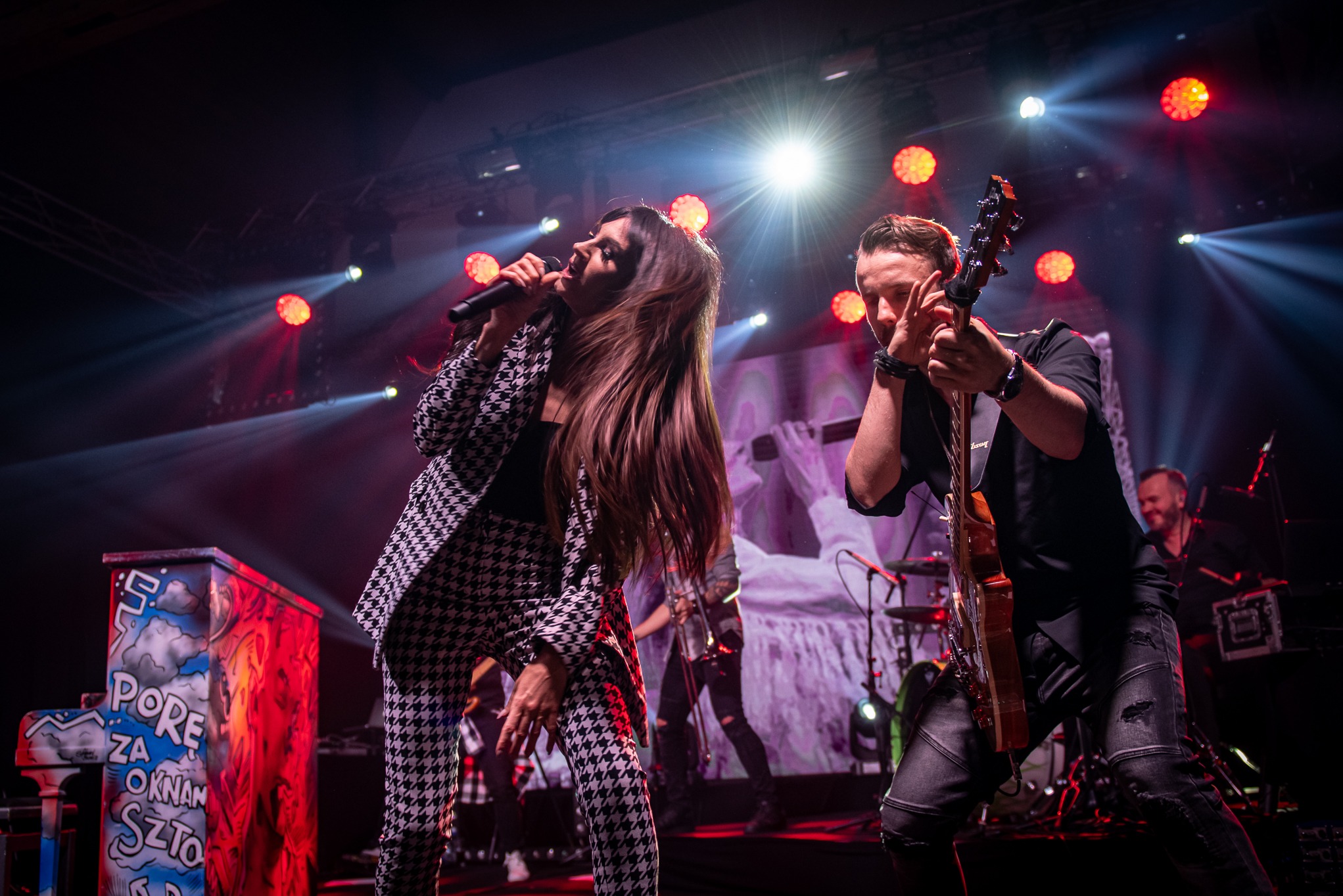 Zespół muzyczny na scenie: wokalistka w kostiumie w pepitkę śpiewa do mikrofonu, gitarzysta gra na gitarze. W tle oświetlenie sceniczne i perkusja.