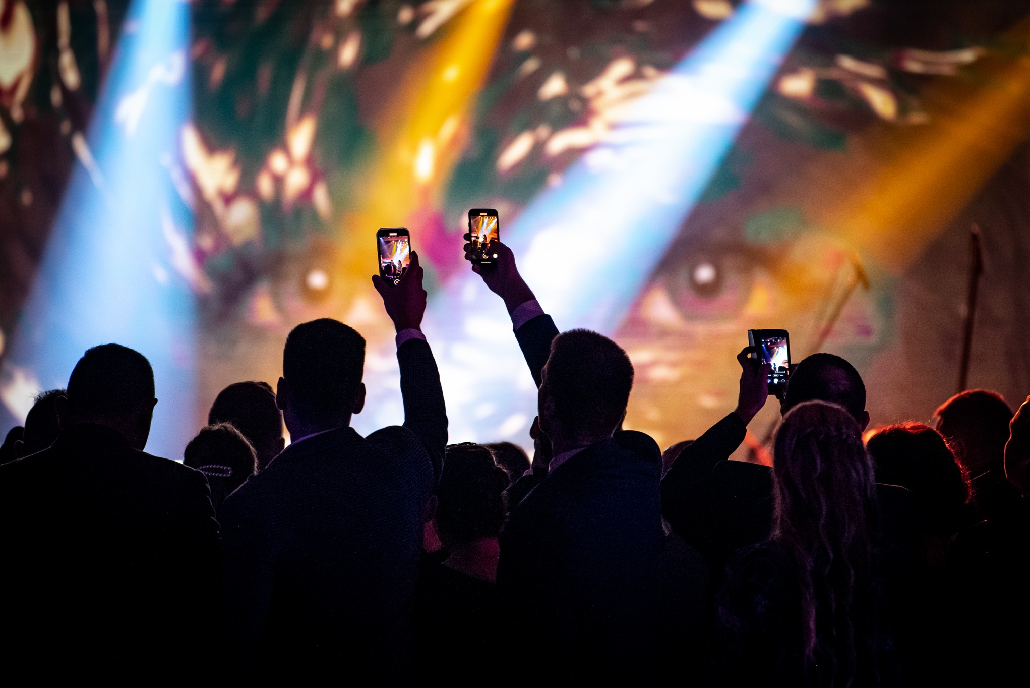 Tłum ludzi filmujących smartfonami scenę z projekcją oka w tle, oświetloną reflektorami podczas eventu.