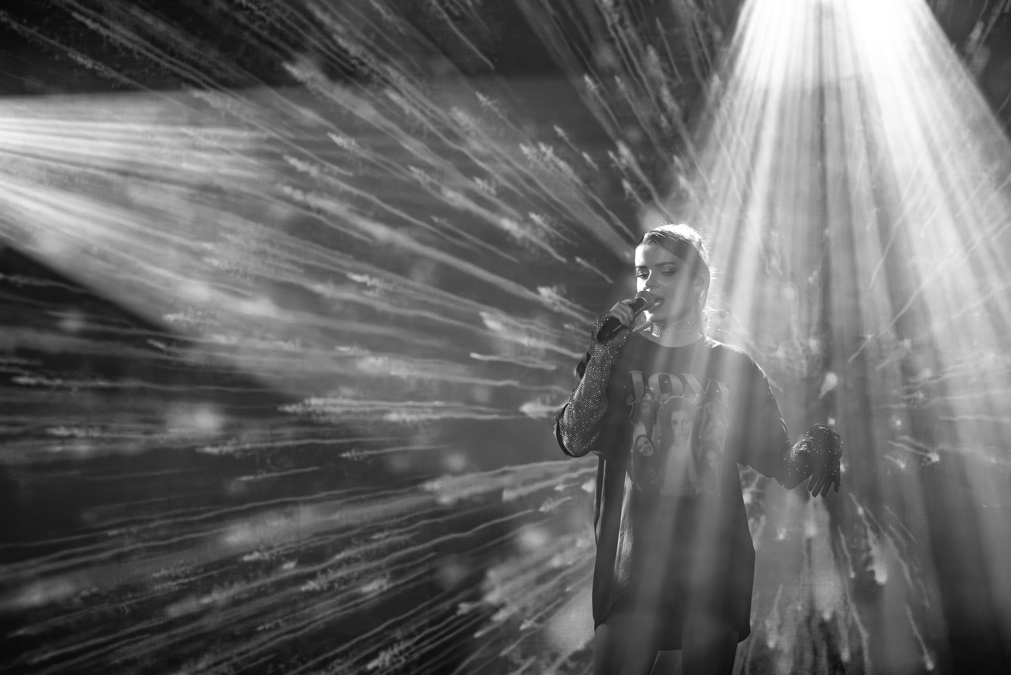 Czarno-białe zdjęcie piosenkarki z mikrofonem na scenie, oświetlonej reflektorami i z abstrakcyjnym tłem, uchwycone z niskiego kąta.