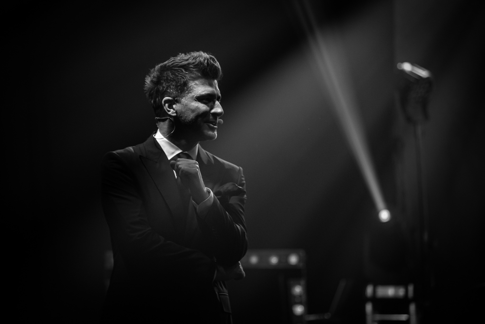 Czarno-białe zdjęcie konferansjera w garniturze, oświetlonego reflektorem, z mikrofonem nagłownym, w trakcie przemówienia na tle ciemnej sceny.