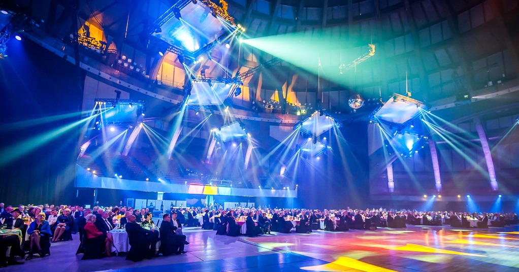 Uroczysta gala w hali widowiskowej: rzędy stolików z gośćmi, oświetlenie sceniczne i dekoracje. Parkiet oświetlony kolorowymi reflektorami.