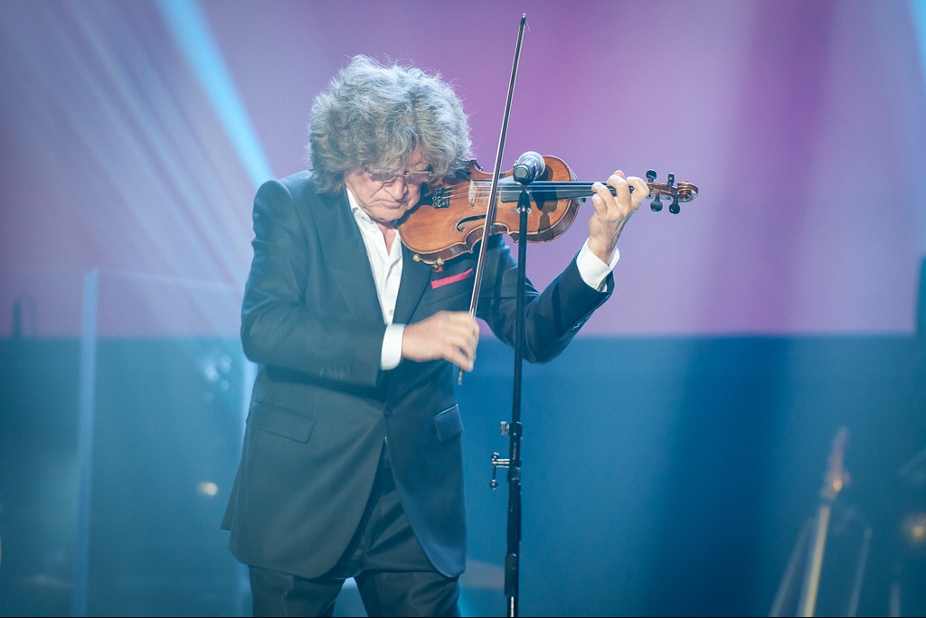 Skupiony skrzypek w garniturze gra na skrzypcach na scenie z oświetleniem eventowym. Widoczny statyw mikrofonowy i fragment gitary w tle.