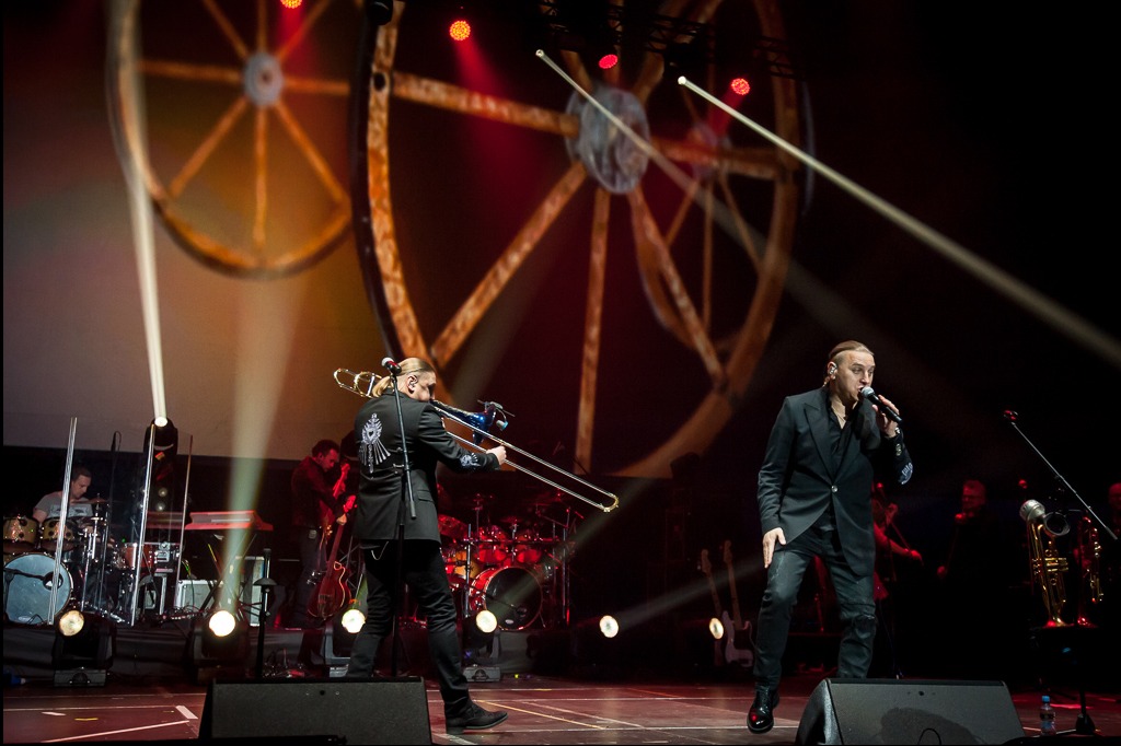 Zespół muzyczny na scenie podczas koncertu: wokalista w czarnym garniturze i trębacz z ozdobną marynarką, w tle perkusja i oświetlenie sceniczne.