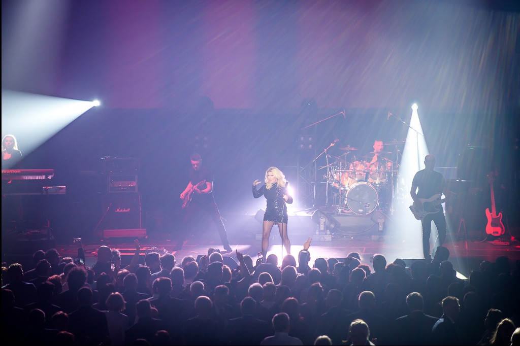 Zespół muzyczny na scenie podczas koncertu. Wokalistka w błyszczącej sukience, gitarzyści i perkusista w tle. Publiczność z podniesionymi rękami. Scena oświetlona reflektorami.