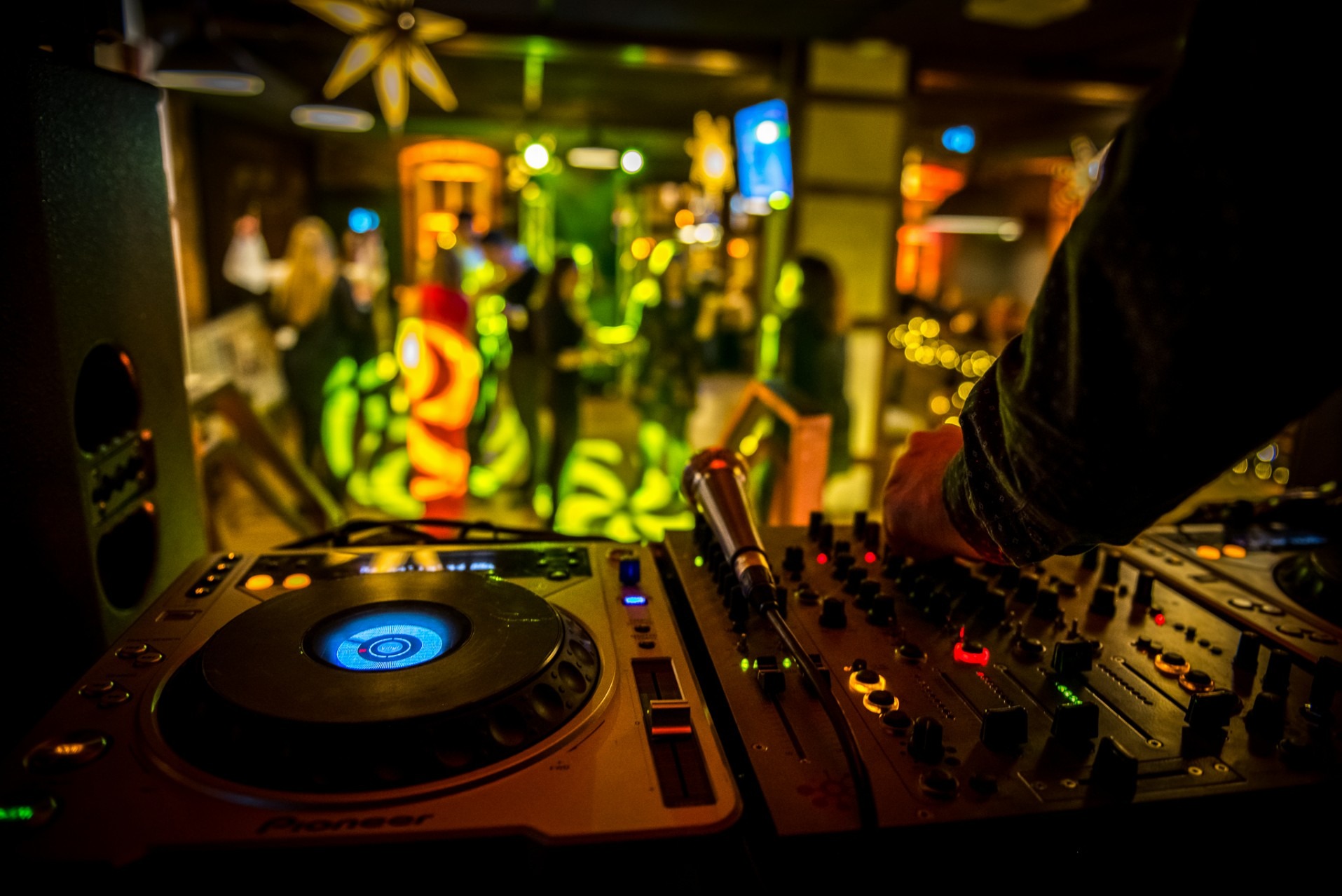 DJ miksuje muzykę na konsolecie Pioneer podczas eventu. W tle rozmyte sylwetki osób i dekoracje świetlne tworzą imprezowy klimat.