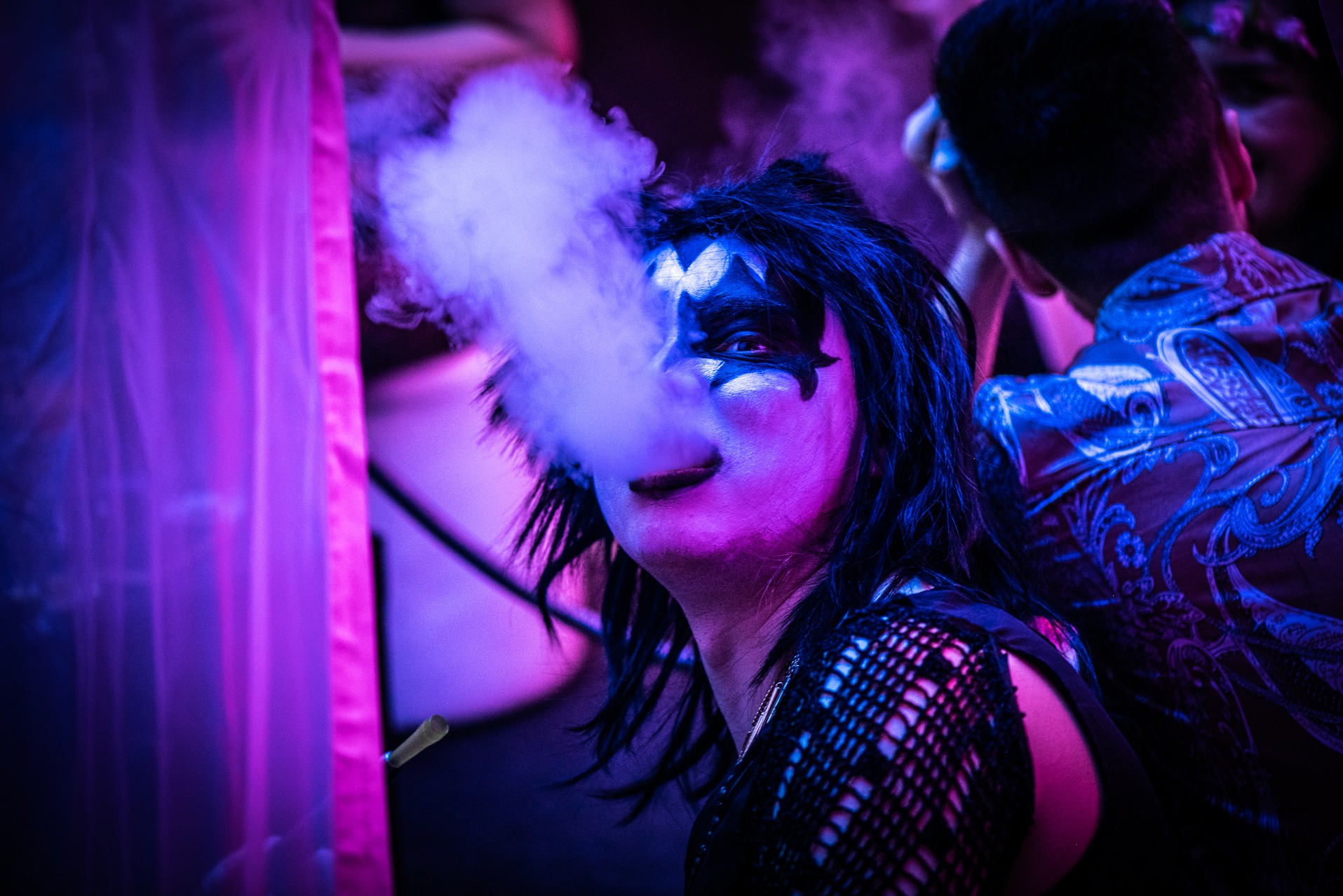 Osoba z makijażem w stylu KISS, wypuszczająca dym, oświetlona fioletowym światłem, widoczna w tle inna osoba w wzorzystej koszuli. Zdjęcie z wydarzenia.