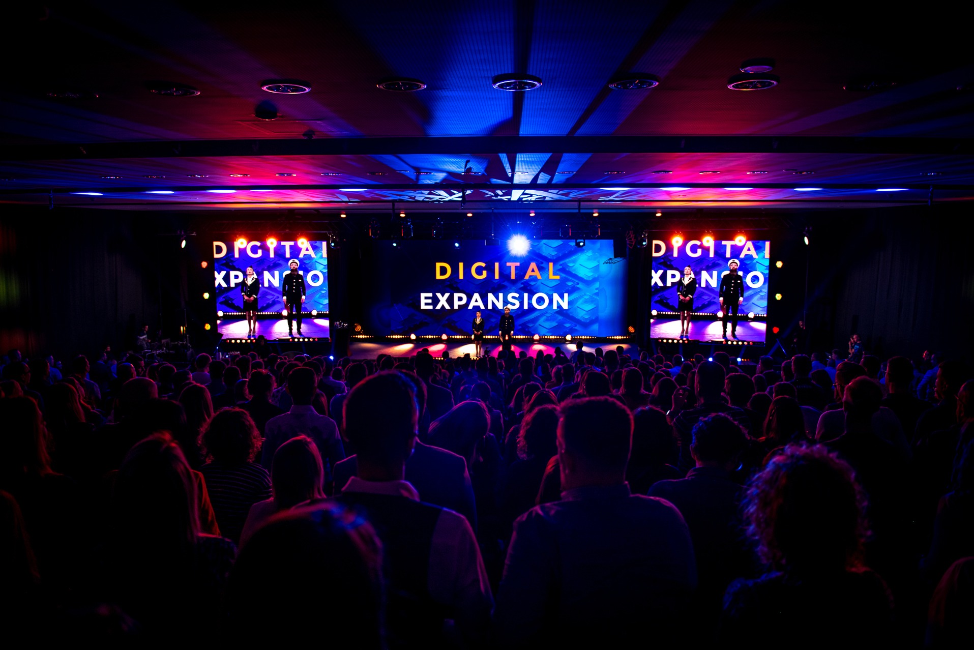 Widok na scenę z publicznością podczas konferencji 'Digital Expansion'. Na scenie osoby w kostiumach marynarskich, ekrany LED i efekty świetlne.