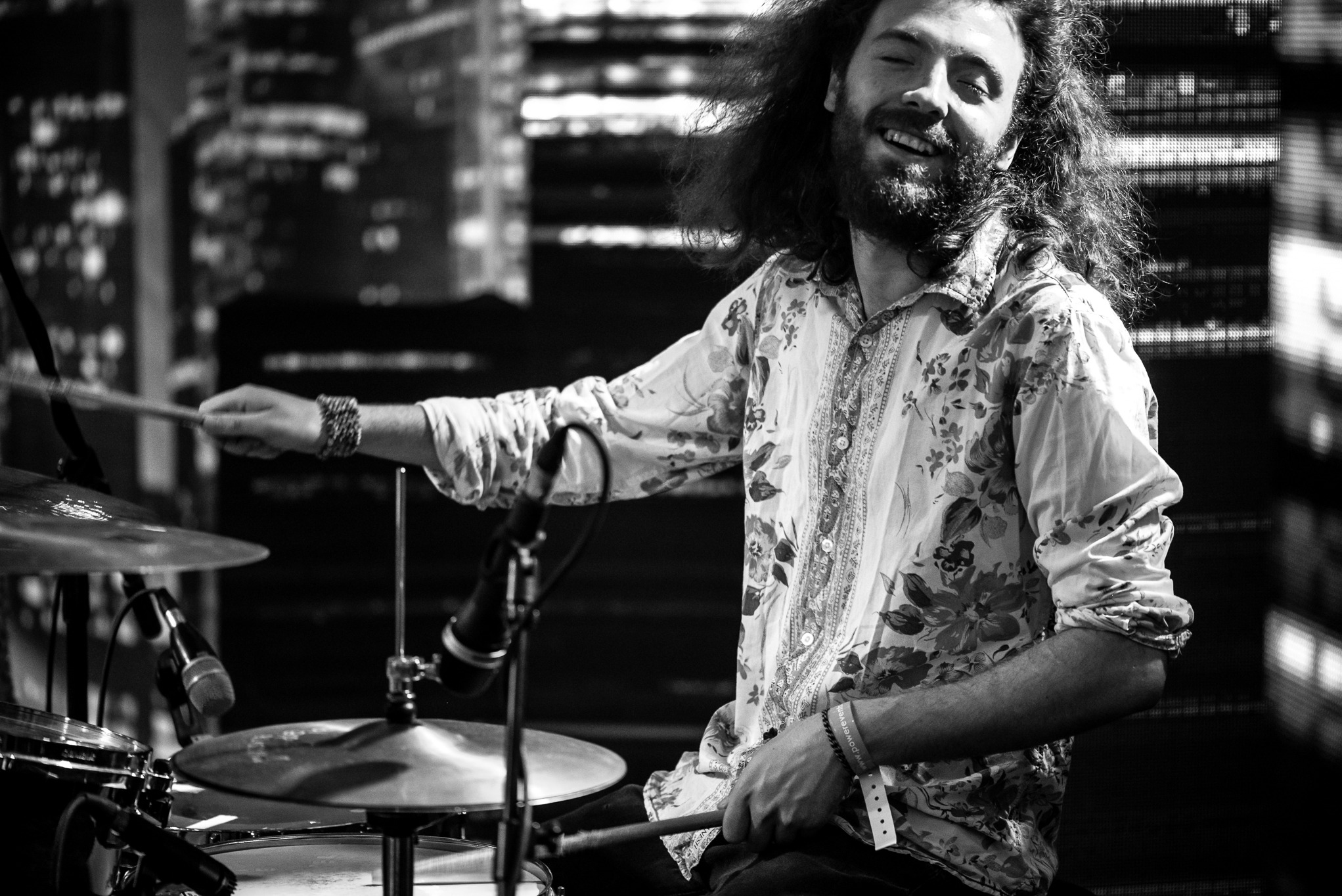 Czarno-białe zdjęcie uśmiechniętego perkusisty z długimi włosami w kwiecistej koszuli, grającego na perkusji podczas koncertu. W tle rozmazane światła miasta.