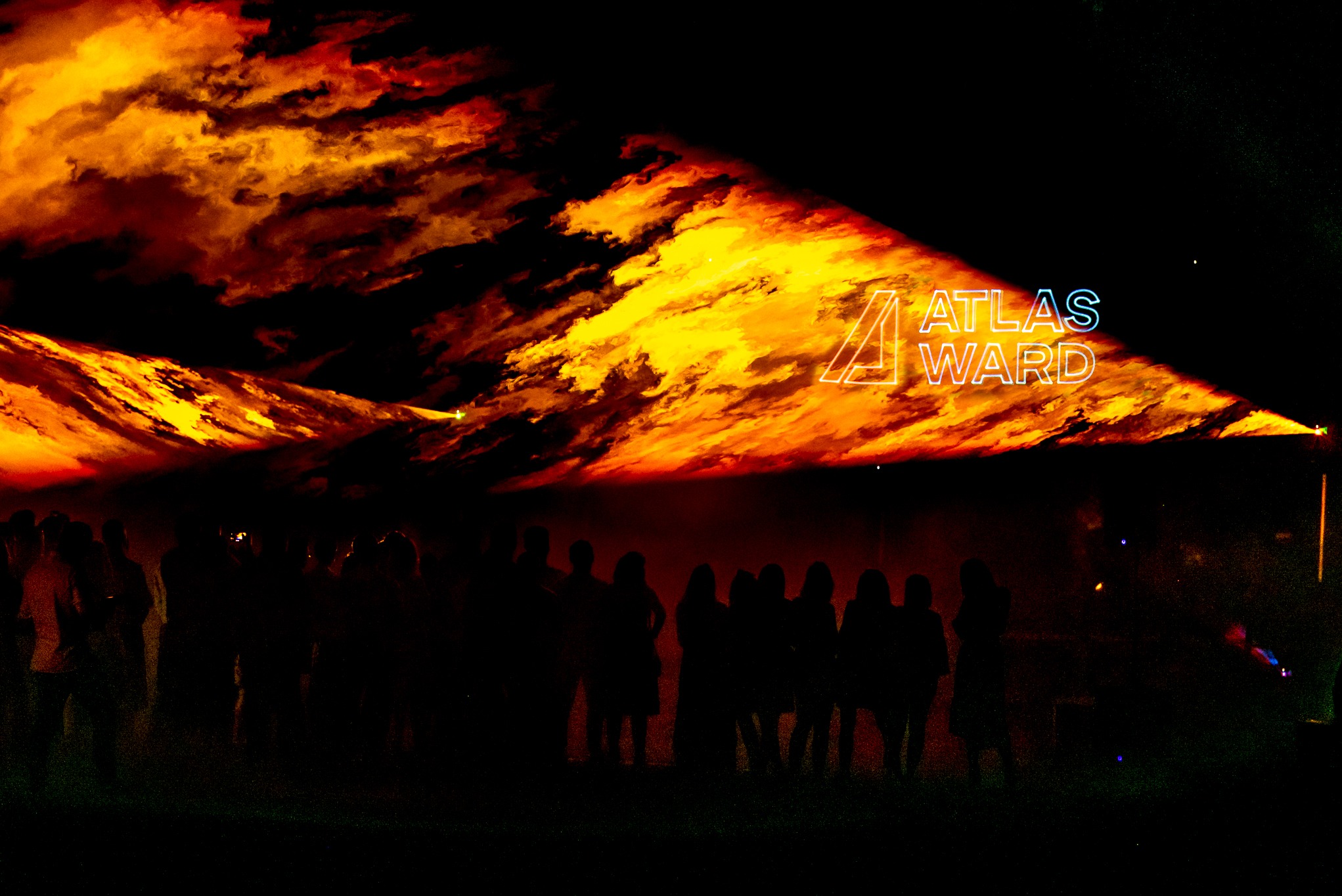 Tłum ludzi w ciemności, oświetlony pomarańczowo-żółtymi światłami i logo 'ATLAS WARD'. Efektowne oświetlenie sceniczne tworzy dramatyczną atmosferę.