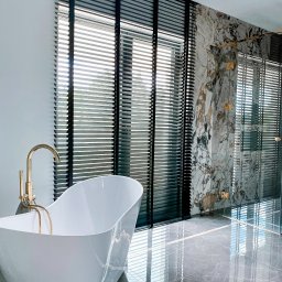 Aurena Marcin Szóstko - Luksusowa łazienka z wolnostojącą wanną, złotą armaturą i ciemnymi żaluzjami poziomymi na oknach oraz w kabinie prysznicowej, marmurowe wykończenie ścian i podłogi.