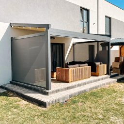 Aurena Marcin Szóstko - Nowoczesny taras z szarą pergolą i boczną osłoną przeciwsłoneczną, meble ogrodowe z drewna tekowego, jasna elewacja budynku w tle.