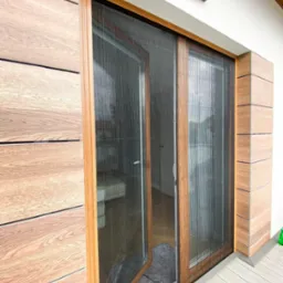 Zamknięta i częściowo otwarta plisowana siatka w drewnianej ramie, chroniąca przed owadami, zamontowana na tarasie z drewnianą elewacją.