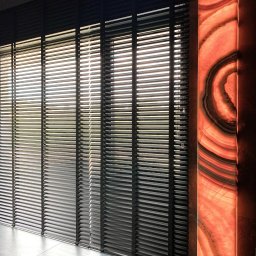 Aurena Marcin Szóstko - Ciemne, poziome osłony okienne z cienkimi lamelami, obok podświetlany panel z agatu w odcieniach pomarańczy i brązu, na podłodze płytki imitujące kamień.