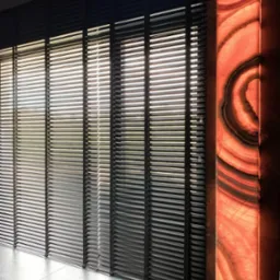 Ciemne, poziome osłony okienne z cienkimi lamelami, obok podświetlany panel z agatu w odcieniach pomarańczy i brązu, na podłodze płytki imitujące kamień.