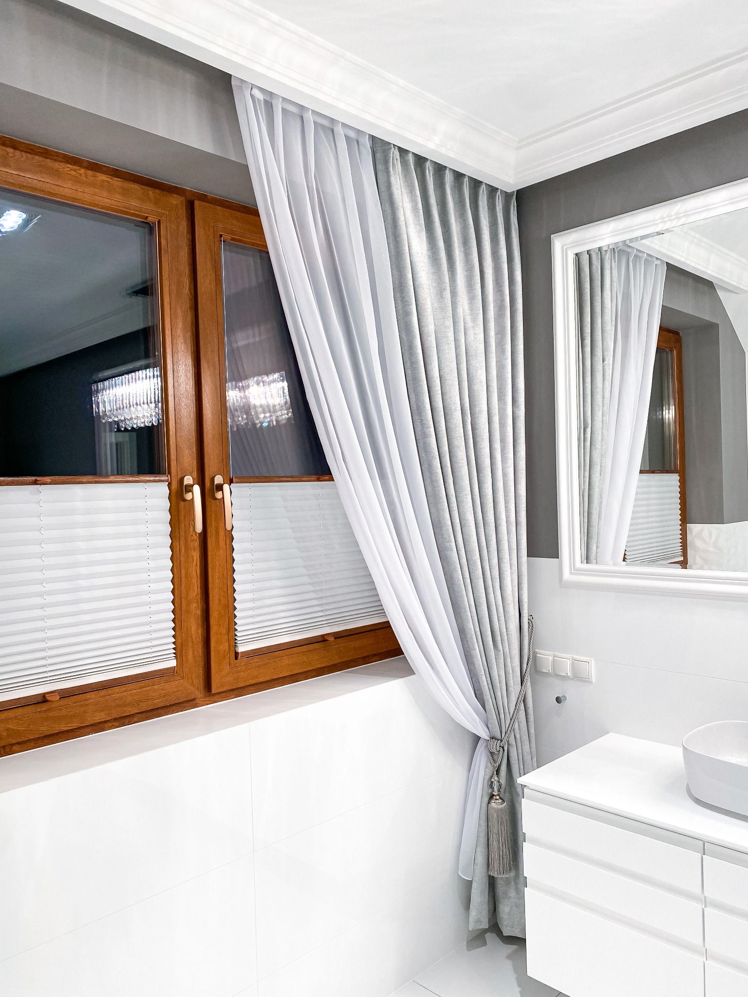 Jasne, plisowane rolety wewnętrzne w drewnianej ramie okiennej, uzupełnione o biało-szare, dekoracyjne zasłony z ozdobnym wiązaniem i frędzlami w łazience z białą umywalką i lustrem.