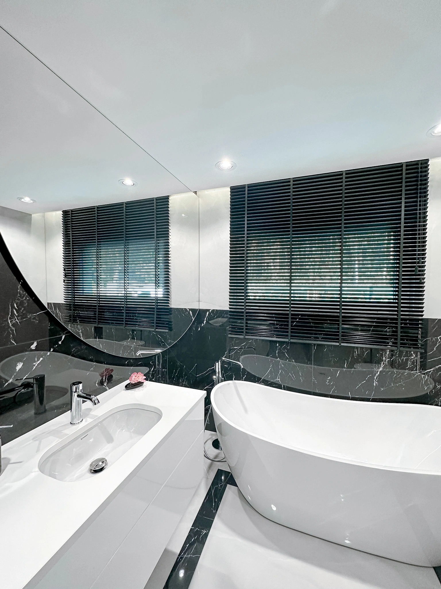 Elegancka łazienka z czarno-białym marmurem, owalną umywalką, wolnostojącą wanną i ciemnymi żaluzjami poziomymi w oknie, odbijającymi się w lustrze.