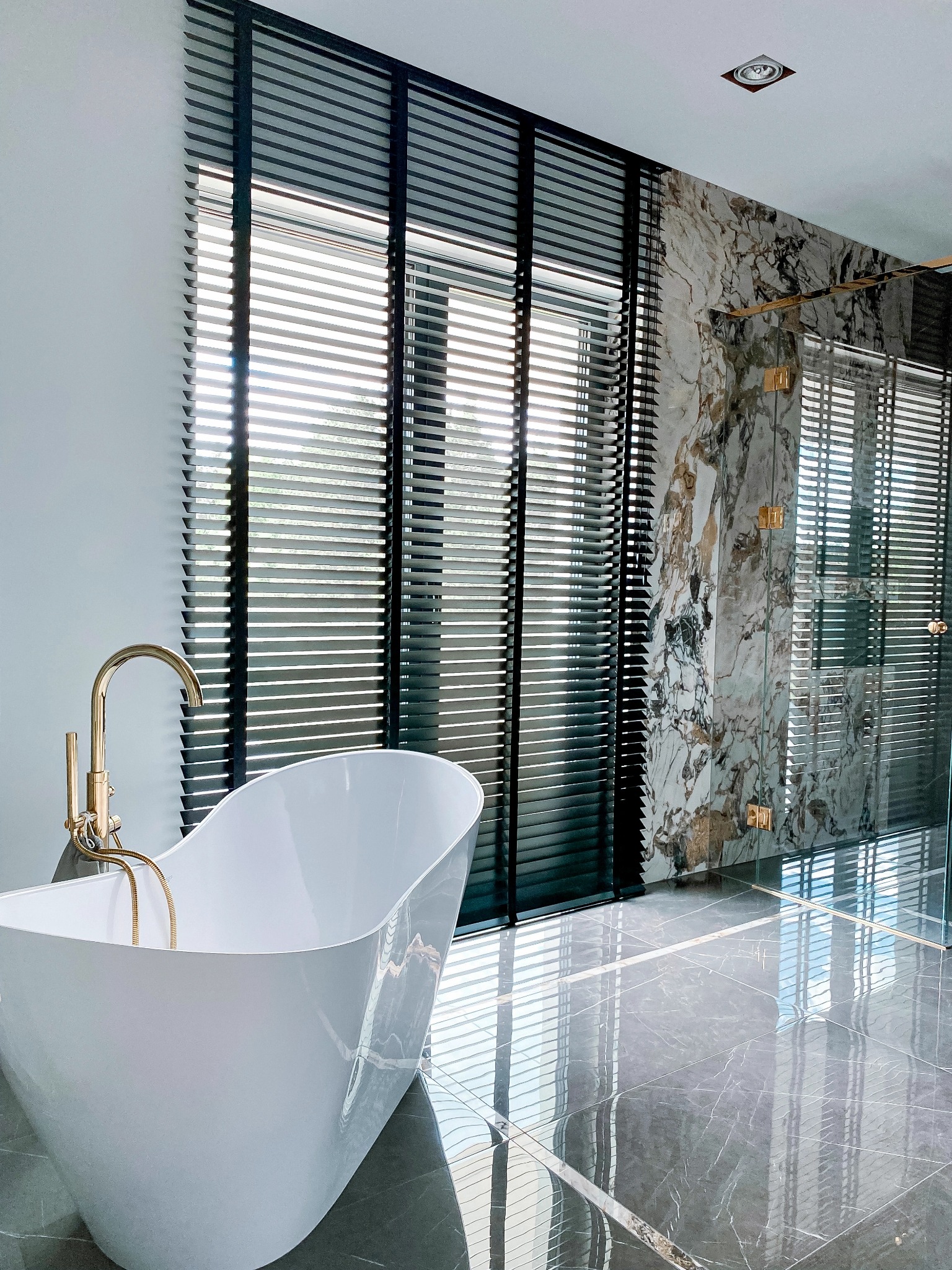 Luksusowa łazienka z wolnostojącą wanną, złotą armaturą i ciemnymi żaluzjami poziomymi na oknach oraz w kabinie prysznicowej, marmurowe wykończenie ścian i podłogi.