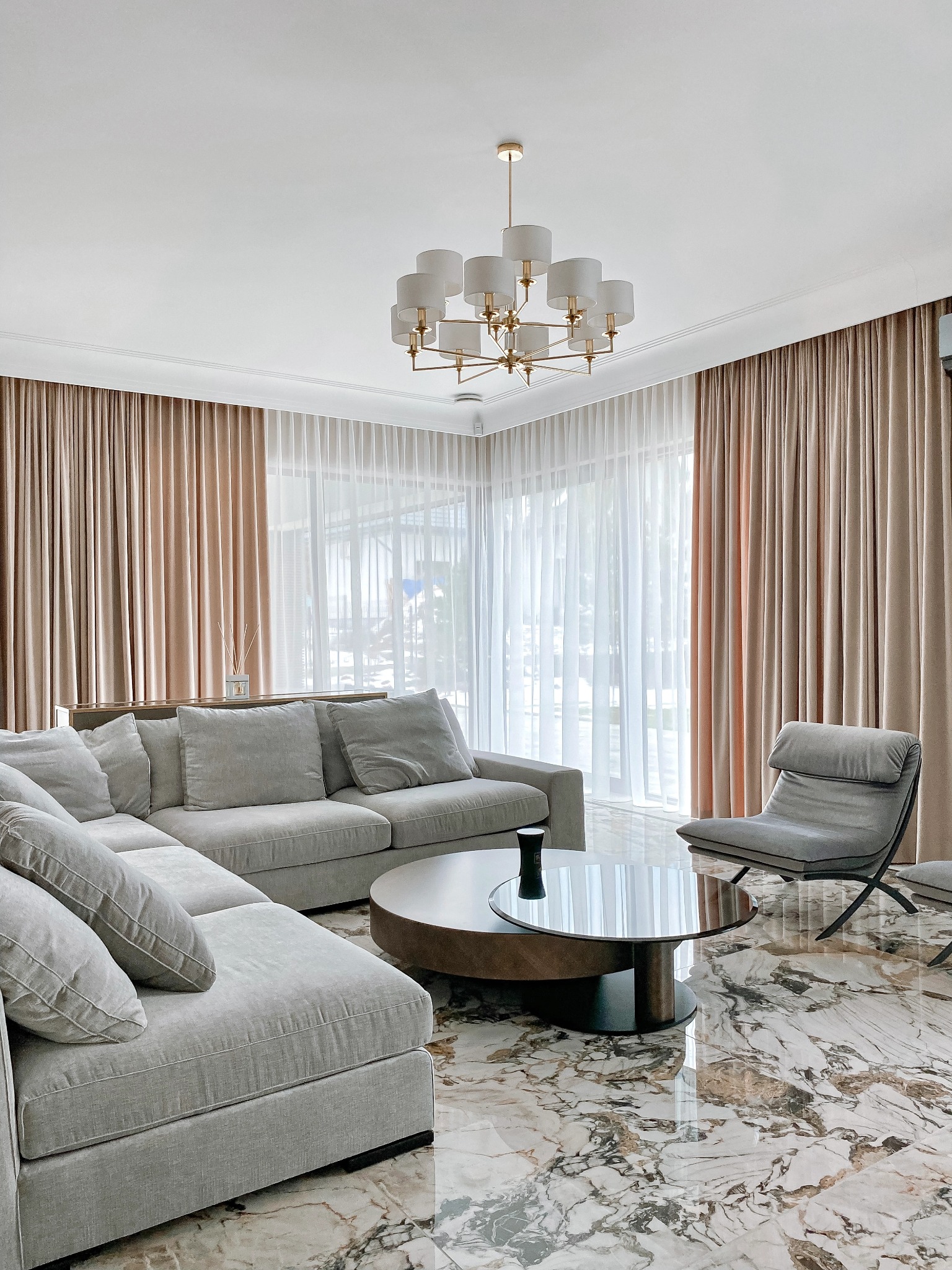 Eleganckie wnętrze salonu z beżowymi zasłonami i firanami, szarą sofą narożną, okrągłym stolikiem kawowym z szklanym blatem i marmurową podłogą.