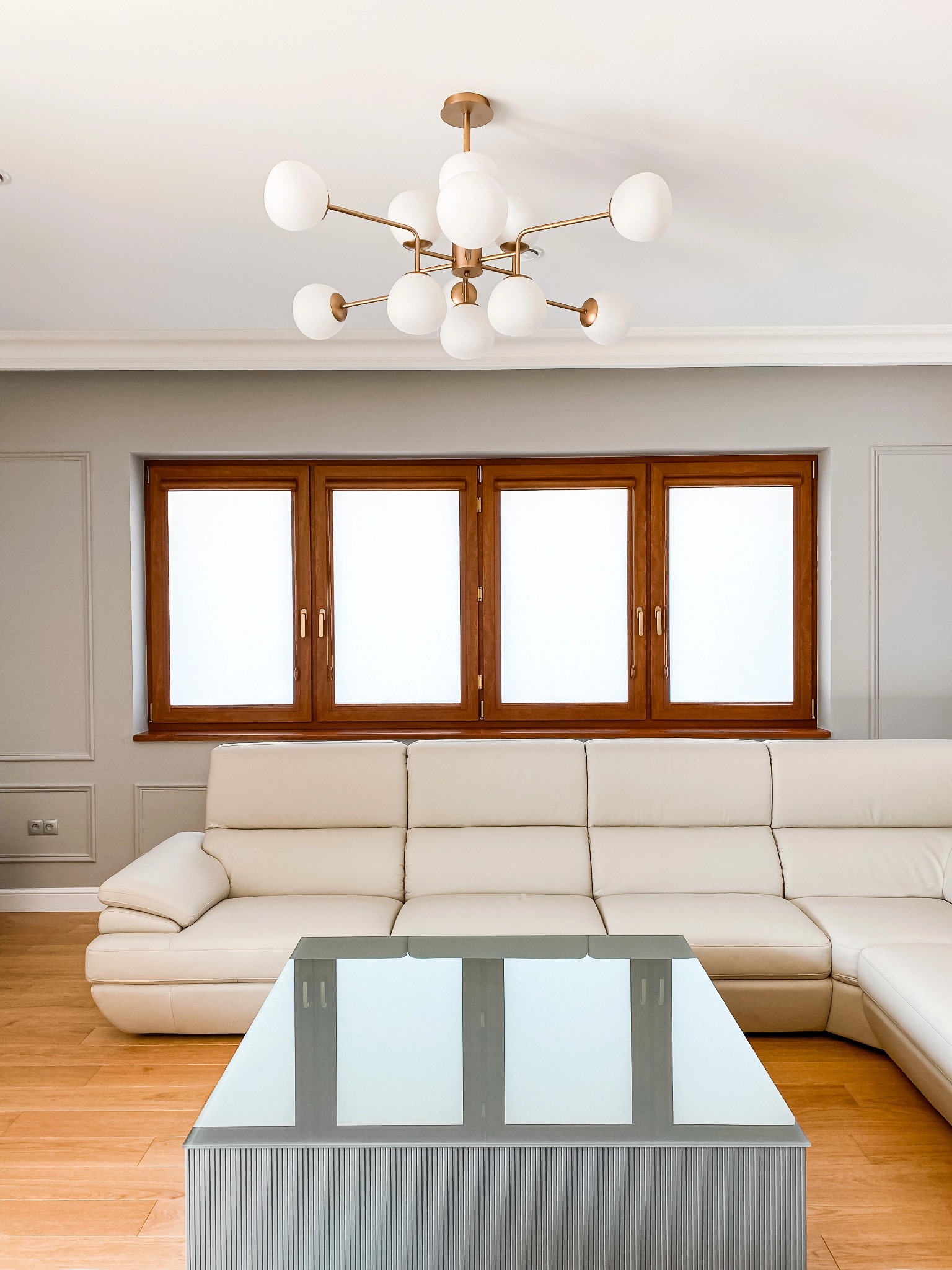 Jasne wnętrze salonu z drewnianymi oknami zasłoniętymi białym materiałem, kremową skórzaną sofą narożną i szklanym stolikiem kawowym na parkiecie.