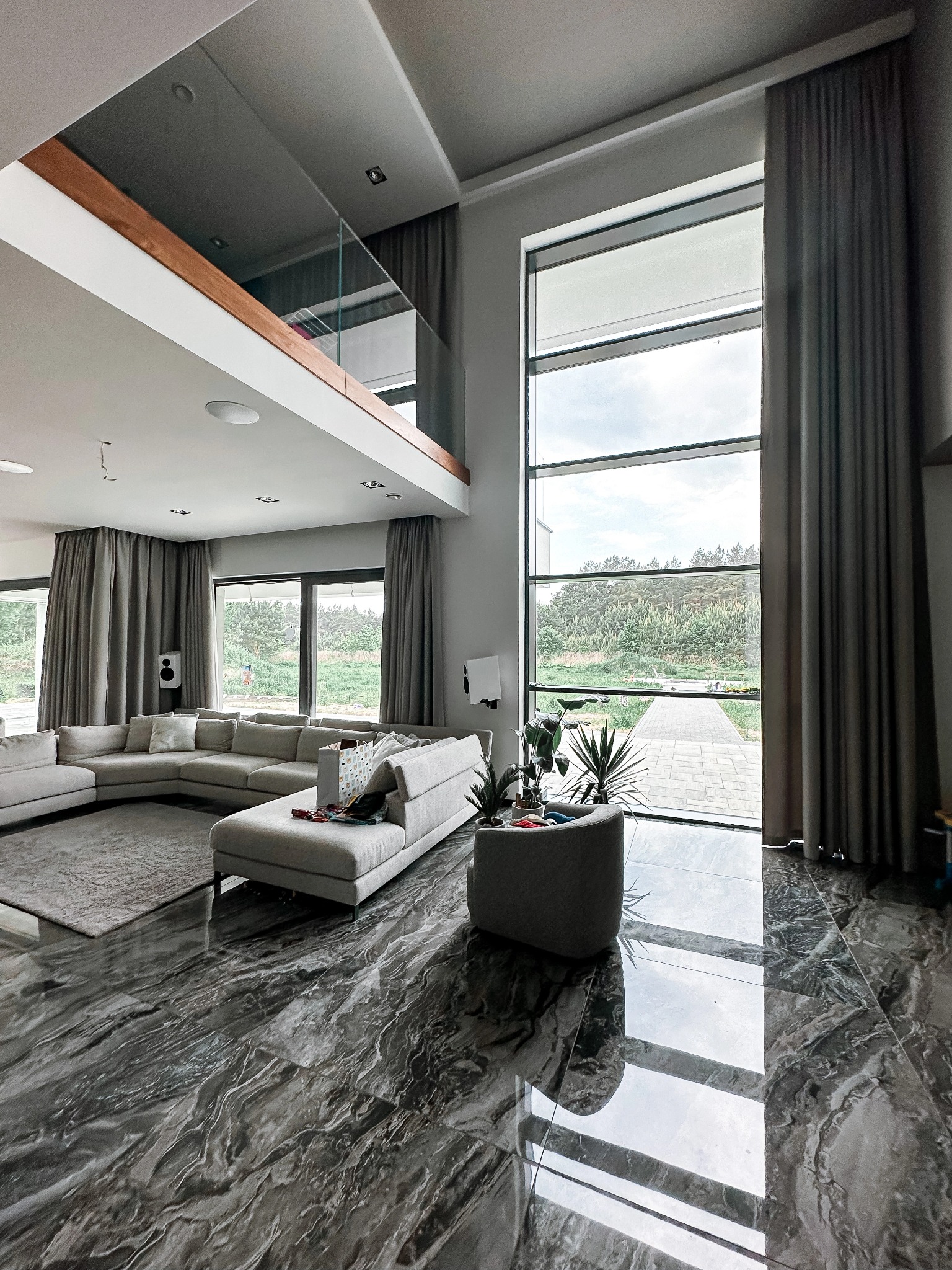 Wnętrze przestronnego salonu w Białymstoku z wysokimi oknami przesłoniętymi grafitowymi zasłonami, marmurowa podłoga odbija światło, widoczny narożnik sofy i antresola z szklaną balustradą.