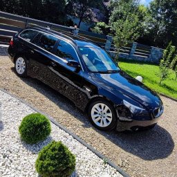 Detailing Manufaktura Blasku Powłoka Ceramiczna, Sprzątanie i Polerowanie Samochodu- - Czarny samochód kombi zaparkowany na białym żwirze, z dwoma kulistymi krzewami i kamienną kulą dekoracyjną w tle, widok na zadbany ogród i błękitne niebo.