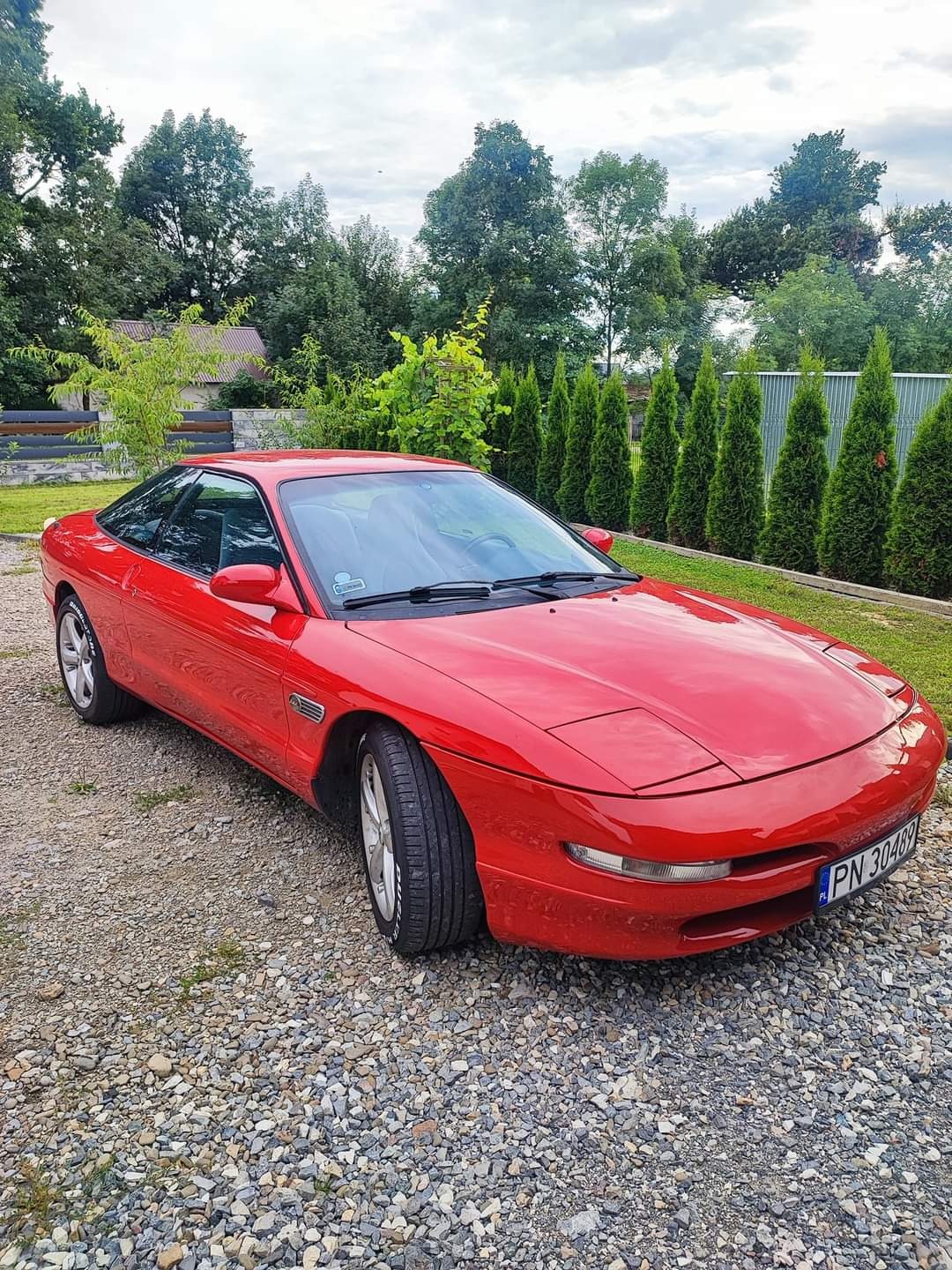 Czerwone sportowe auto Ford Probe zaparkowane na żwirowym podjeździe, widoczne tablice rejestracyjne, w tle żywopłot z iglaków i drzewa.