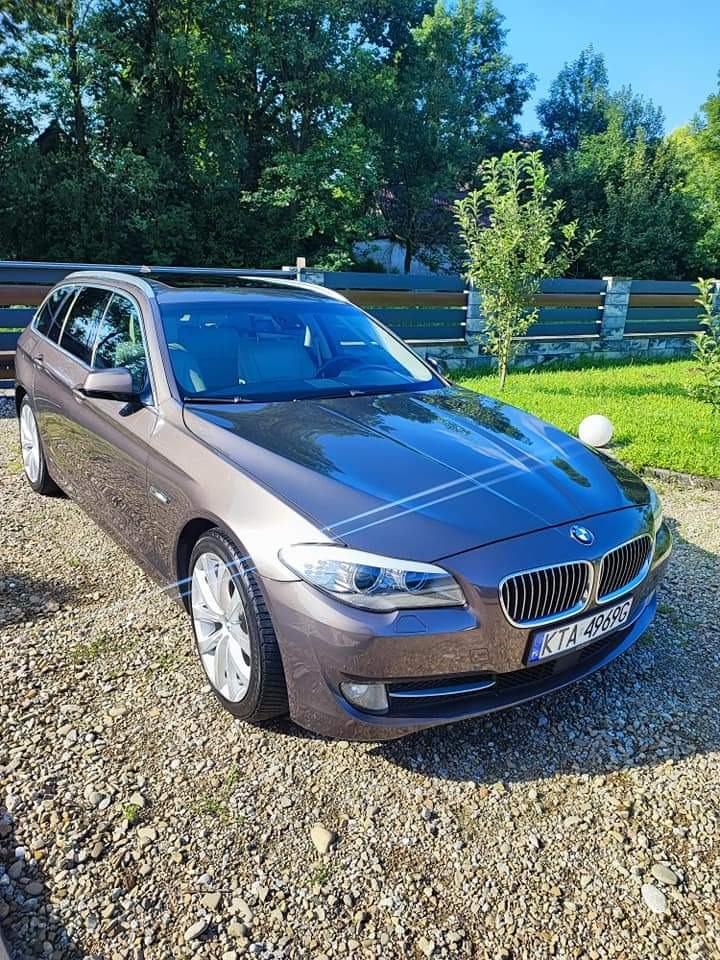 Czysty, brązowy samochód BMW serii 5 kombi zaparkowany na żwirowym podjeździe, z widoczną tablicą rejestracyjną KTA 49696, na tle zieleni drzew i ogrodzenia.