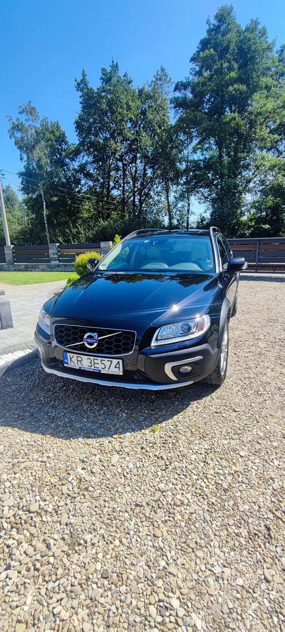 Błyszczący, czarny Volvo XC70 zaparkowany na żwirowym podjeździe w słoneczny dzień, z widoczną tablicą rejestracyjną KR 3E574 i zielonym tłem drzew.