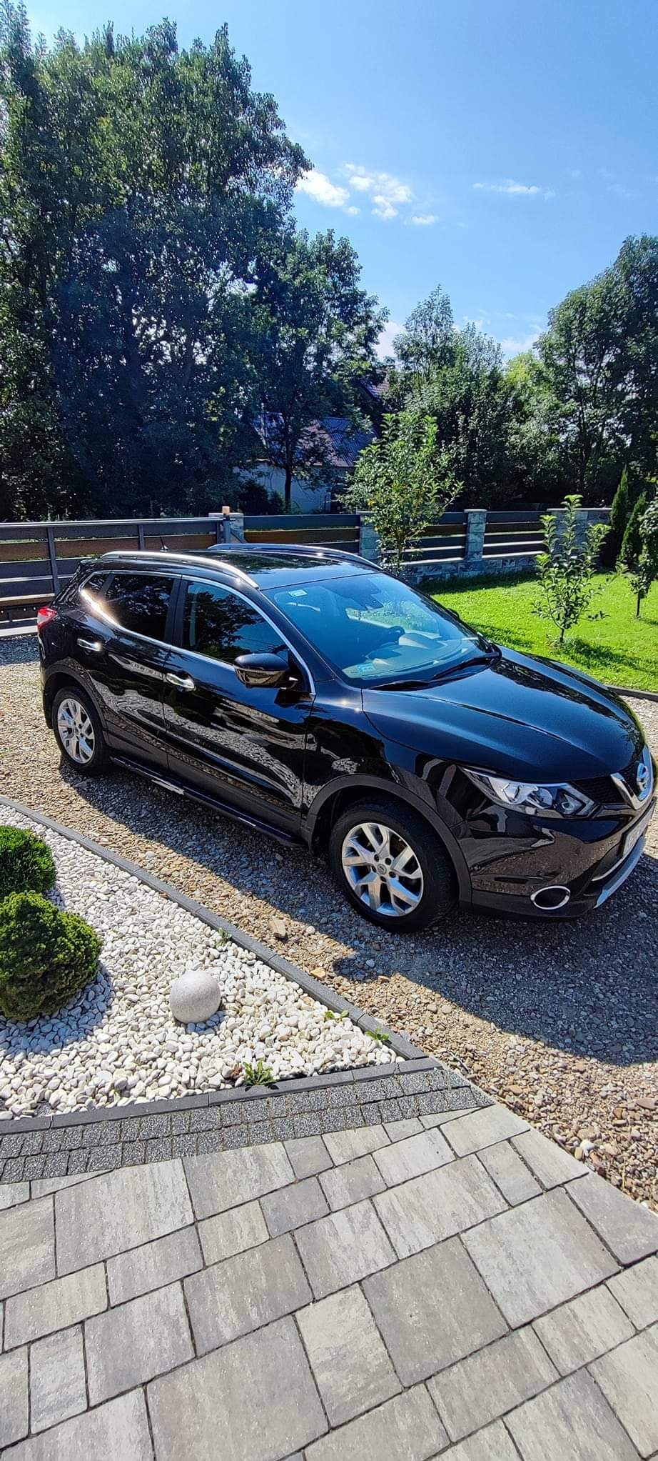 Błyszczący, czarny Nissan Qashqai zaparkowany na żwirowym podjeździe z kostki, w tle zielony trawnik i drewniany płot.